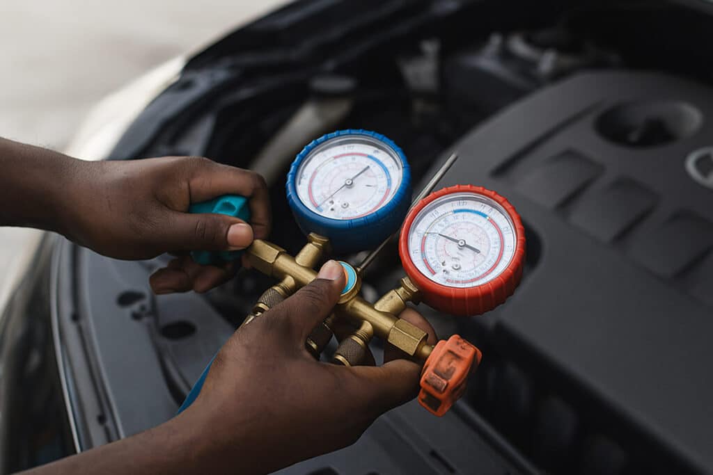 A imagem mostra um mecânico operando um manômetro, uma ferramenta utilizada para verificar e recarregar o sistema de ar condicionado automotivo.