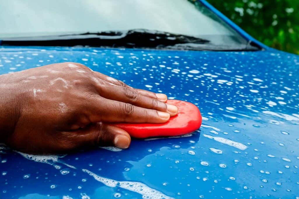 A imagem mostra uma pessoa utilizando uma Clay Bar (também conhecida como barra descontaminante) em um carro azul, que está coberto por espuma e água.