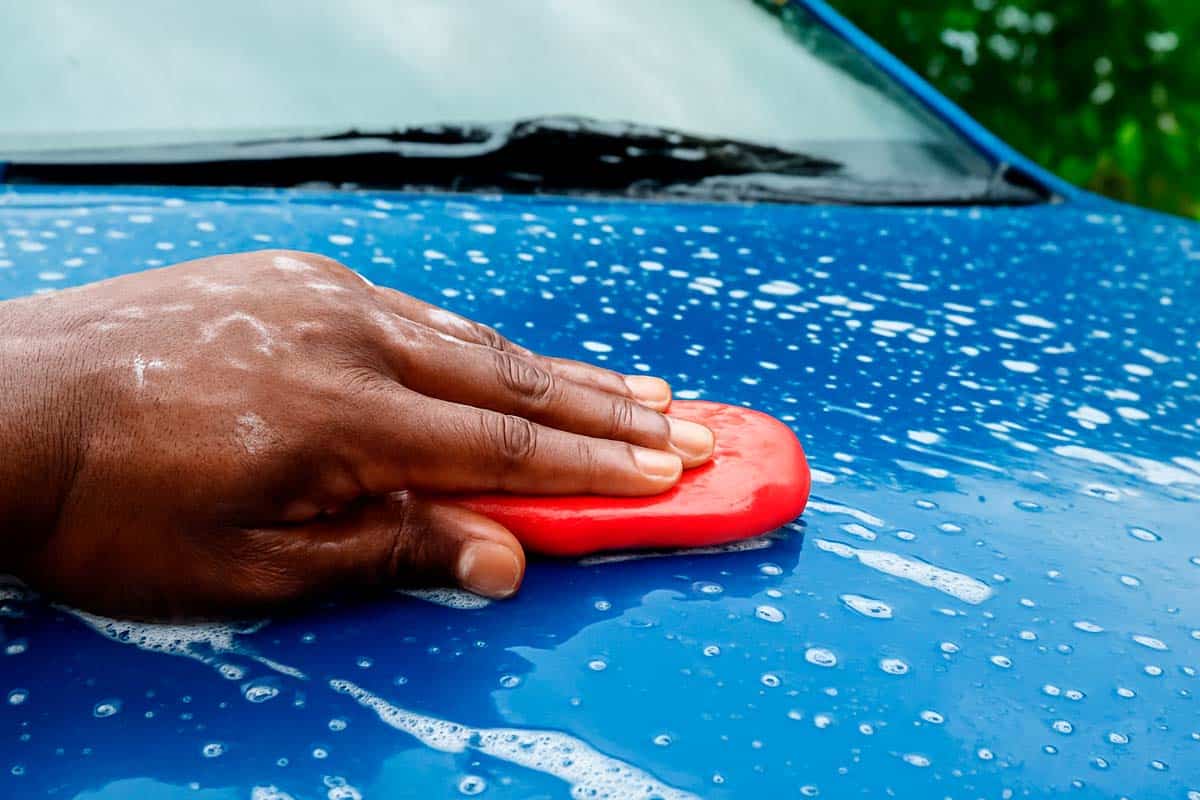 A imagem mostra uma pessoa utilizando uma Clay Bar (também conhecida como barra descontaminante) em um carro azul, que está coberto por espuma e água.